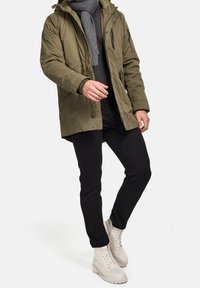 Olivegrüner Parka mit Kapuze, Reißverschluss und Knopfverschlüssen, kombiniert mit schwarzen, schmal geschnittenen Hosen und beigefarbenen Schnürboots. Grauer Schal.