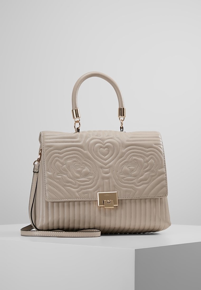 Sac à main en cuir beige avec motifs floraux et cœurs matelassés, fermoir doré, poignée supérieure et bandoulière amovible sur une surface blanche.