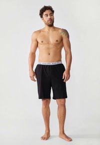 Uomo senza maglietta con un tatuaggio sulla spalla sinistra, indossando pantaloni corti neri con una cintura bianca con la scritta "PIER ONE", in piedi a piedi nudi su uno sfondo chiaro.