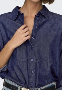 Person wearing a dark blue denim button-up shirt tucked into light blue jeans with a black belt, hand adjusting shirt collar.
