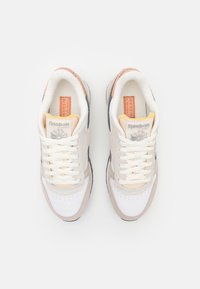 Zapatillas deportivas beige y blancas con material de malla y ante, agujetas planas, marca Reebok y suela negra. Acentos naranjas en el talón.