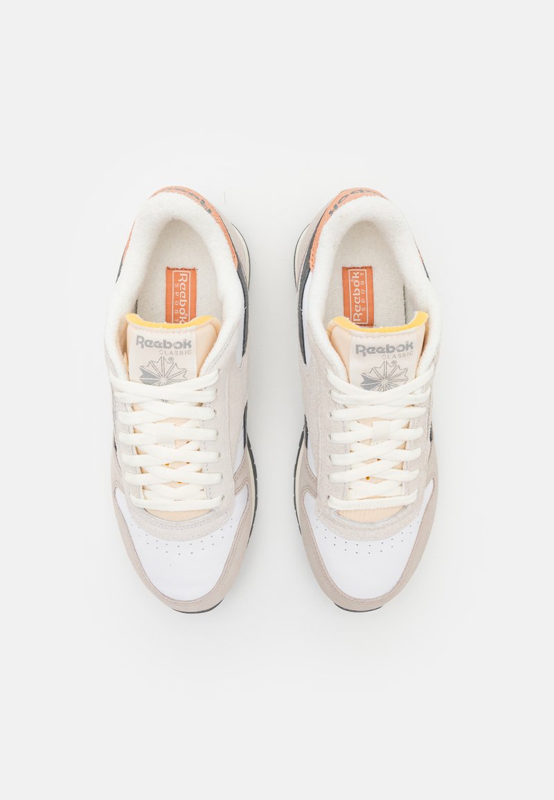 Baskets de sport beige et blanches avec des matériaux en maille et en daim, lacets plats, marquage Reebok et semelle extérieure noire. Accents oranges sur le talon.