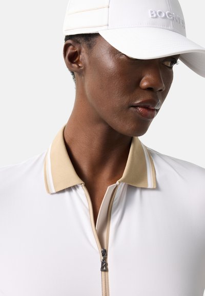 Femme portant un polo blanc zippé avec un col beige et une casquette blanche brodée du mot « Bogn », regardant sur le côté.