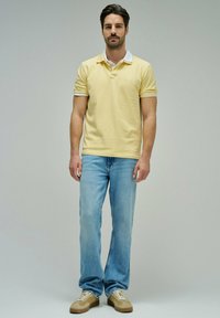 Hombre de pie, de frente, vistiendo una camisa polo amarilla pálida, jeans azul claro y zapatillas beige, sobre un fondo liso.