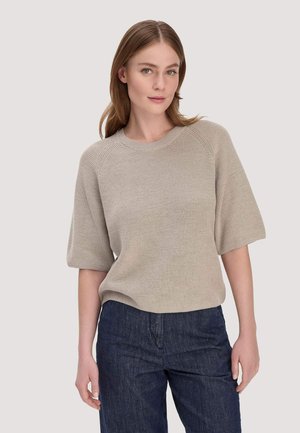 Junge Frau mit hellbraunen Haaren, die einen beige kurzärmeligen Strickpullover und dunkelblaue Jeans trägt, vor einem schlichten Hintergrund stehend.