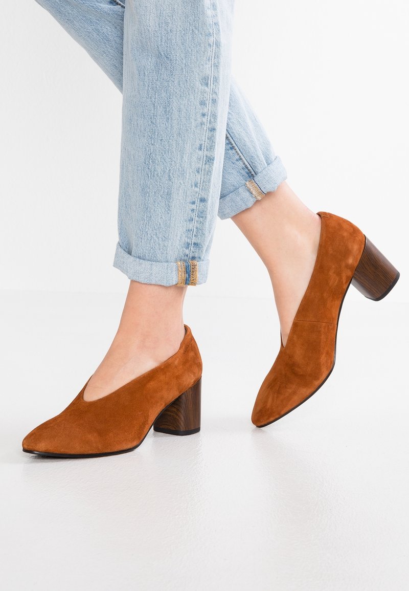 Chaussures à talons en daim marron avec un bout pointu et un design ouvert, présentant un talon en bois texturé et un jean léger bleu retroussé.