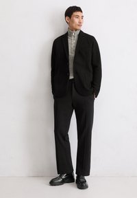 Schwarzer, taillierter Blazer über einem hellgrauen Sweatshirt mit Reißverschluss, kombiniert mit schwarzen Hosen und klobigen schwarzen Schuhen. Glatte Texturen, minimalistisches Design.