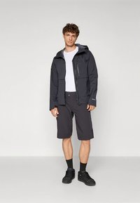 Mladic u crnoj dukserici s kapuljačom, bijeloj majici, crnim šortsevima, crnim čarapama i crnim patikama, stoji naslonjen na svijetlosivu pozadinu.