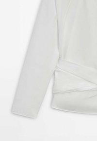 Top blanc à enrouler présentant une texture lisse, des manches longues et un ourlet asymétrique. Le design inclut un détail de nouage sur le côté pour la fermeture.