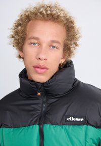 Chaqueta tipo puffer con la parte superior negra y la sección inferior verde, textura suave, alto cuello, cremallera frontal y un logo de ellesse en el pecho.