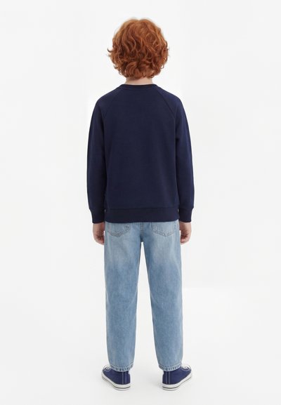 Kind mit lockigem roten Haar steht mit dem Rücken zur Kamera, trägt einen dunkelblauen Pullover, hellblaue Jeans und dunkelblaue Sneakers vor weißem Hintergrund.