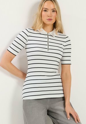 Femme portant un haut blanc côtelé à manches courtes avec des rayures horizontales noires et un col zippé à l'avant, associé à un pantalon gris.