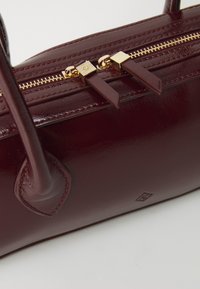 Sac à main en cuir bordeaux avec des fermetures éclair dorées, des poignées doubles et des coutures décoratives. Présente des accessoires carrés et une finition lisse et brillante.