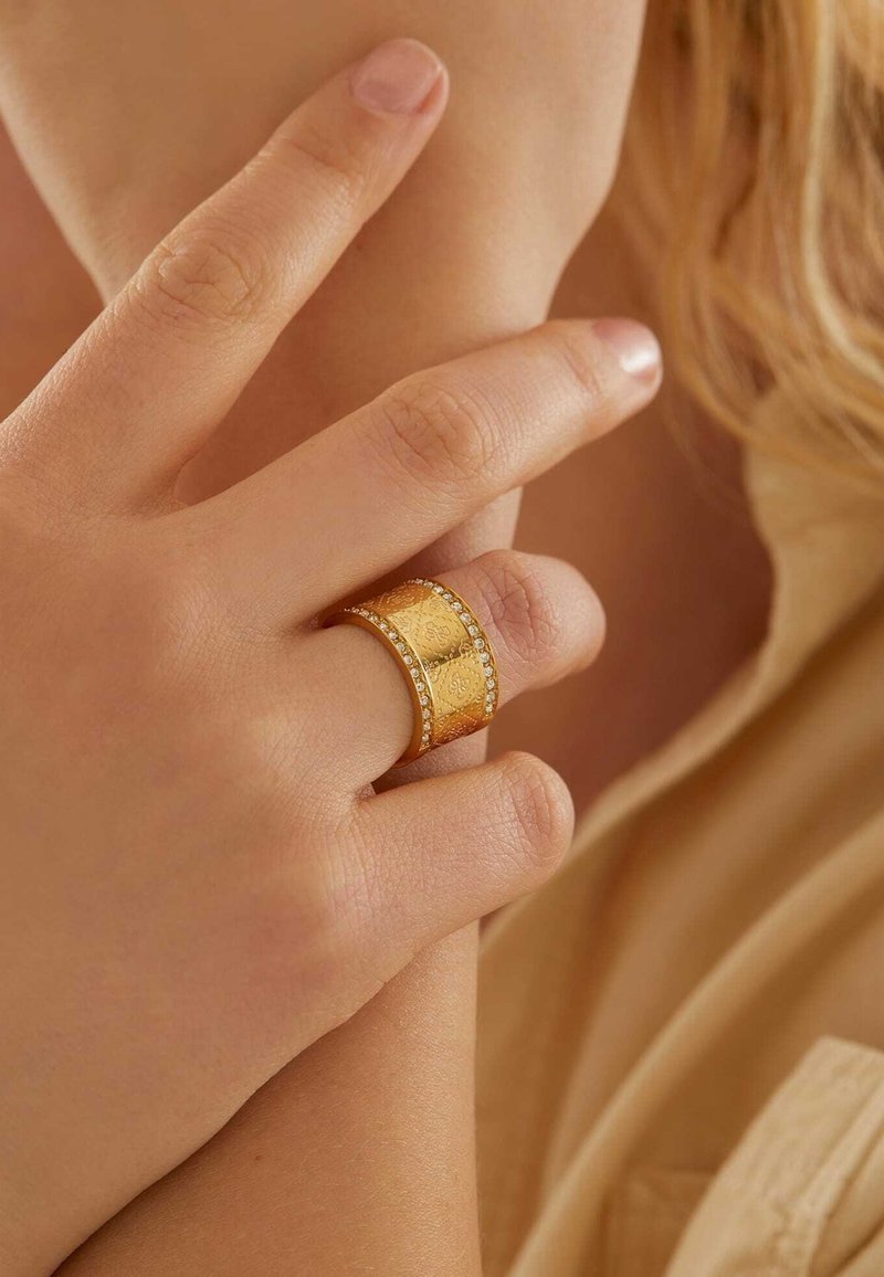 Main avec une large bague en or ornée de motifs gravés et de petits diamants, touchant doucement le bras opposé sur un vêtement beige.