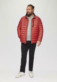 Giacca puffer rossa con tasca sul petto con zip, maglione grigio sotto, pantaloni neri e sneaker dai toni neutri. Texture liscia e design trapuntato.