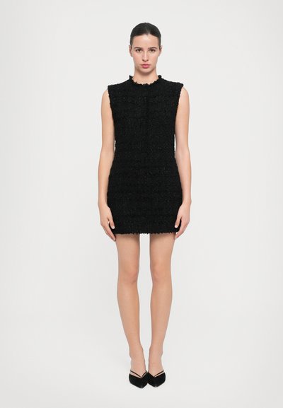 Schwarzes, strukturiertes Kleid mit figurbetonter Silhouette, ärmellosem Design und gewelltem Saum. Akzentuiert mit einem subtilen Schimmer, getragen mit spitzen schwarzen Absätzen.