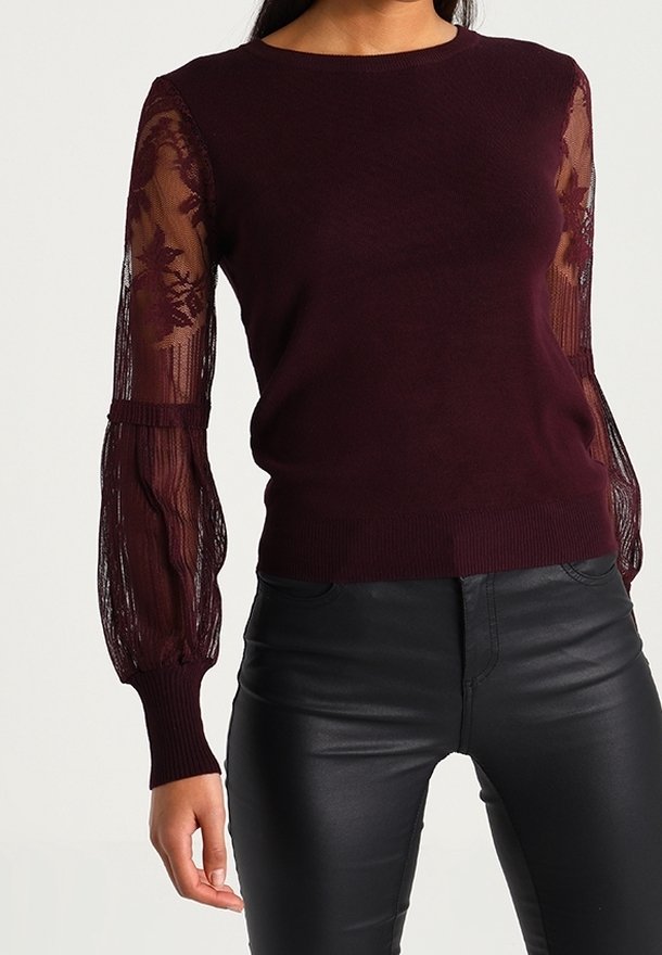 Femme portant un pull violet foncé avec des manches ballon en dentelle transparente et un pantalon en cuir noir ajusté, debout devant un fond clair uni.