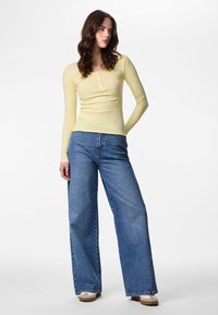 Kvinde står iført en tætsiddende, lysegul top med lange ærmer, højtaljede, vidde ben blå jeans og beige sneakers på en hvid baggrund.