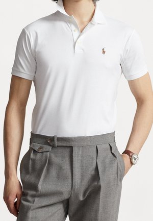 Homme portant un polo blanc à manches courtes avec logo brodé, un pantalon plissé gris et une montre-bracelet avec un bracelet en cuir marron.