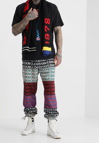 Schwarzes T-Shirt mit Grafikdruck, mehrfarbige gemusterte Hose mit dem Schriftzug "BRAVES" und weiße High-Top-Sneaker. Schwarzer und roter Schal.