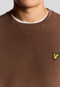 Barna, texturált szövetből készült, bordázott mandzsettákkal és nyakkivágással rendelkező pulóver. A mellrészen kis sárga hímzett logó található.