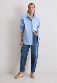 Jasnoniebieska, oversize koszula z guzikami, z kieszenią na piersi, w zestawie z luźnymi, niebieskimi dżinsami i beżowymi trampkami. Proste, czyste linie i swobodny styl.