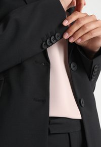 Černý blazer se strukturovaným designem, který má na manžetách čtyři kulaté černé knoflíky a pod ním je vidět světle růžový top.