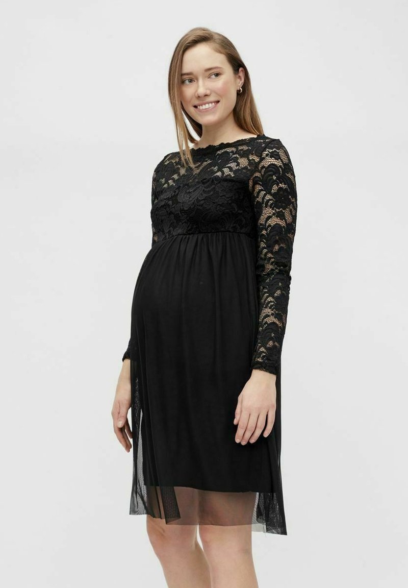Robe de maternité noire avec de longues manches en dentelle, un corsage en dentelle ajusté et une jupe fluide en tissu léger. Détail transparent sur l'ourlet inférieur.