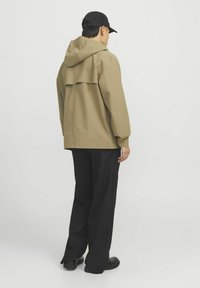 Beige hoodie met een losse pasvorm, voorzien van een rugflapdesign. Gekleed met zwarte broeken en zwarte schoenen, tegen een witte achtergrond.