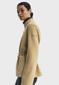 Veste en polaire beige avec col montant, taille à cordon de serrage, poches latérales et détail de logo sur la manche. Texture douce et coupe décontractée.