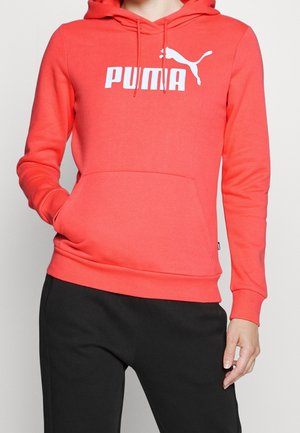Person wearing a bright coral Puma hoodie with front pocket and black pants, hand tucked in pocket, visible from neck to hips.