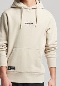 Beige mikina s kapucí, šňůrkou, klokaní kapsou, žebrovanými manžetami a malým logem "SUPERDRY" na hrudi, vyrobená z měkkého materiálu.