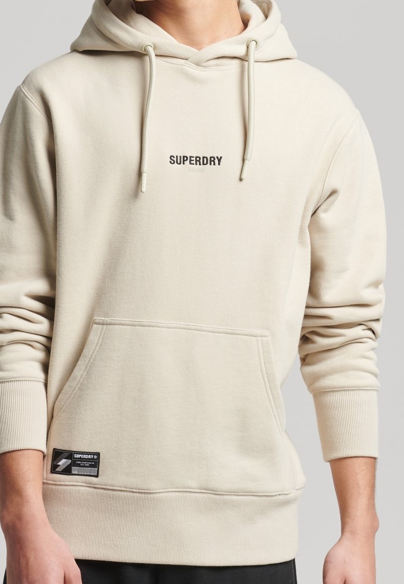 Beige mikina s kapucí, šňůrkou, klokaní kapsou, žebrovanými manžetami a malým logem "SUPERDRY" na hrudi, vyrobená z měkkého materiálu.