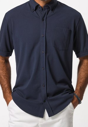 Mann trägt marineblaues kurzärmeliges Hemd mit Knöpfen und Brusttasche sowie weiße Hose, Hände in den Taschen, vor weißem Hintergrund.