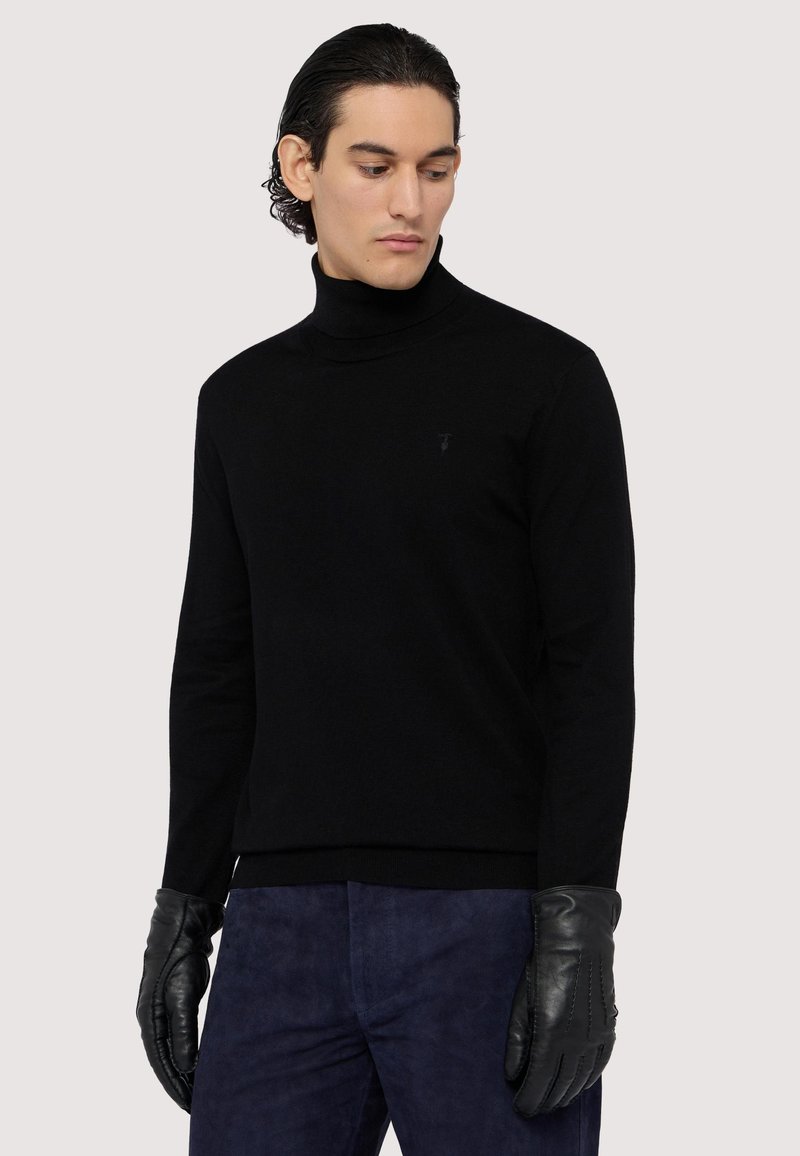 Svart turtleneck-sweater i mjukt material med en subtil logotypdetalj. Matchad med svarta läderhandskar och marinblå strukturerade byxor.