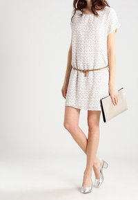 Krátký rukáv, bílá šaty s tečkovaným vzorem, stažené v pase tenkým hnědým páskem; v kombinaci se stříbrnými podpatky a stříbrnou kabelkou.