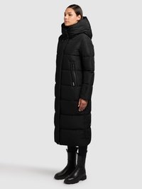 Juoda ilga pūkinė striukė su gobtuvu, pasižyminti dygsniuotu raštu, užtrauktukais užsegamomis kišenėmis ir lieknu siluetu, derinama su juodais odiniais batais.