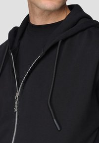 Felpa nera con zip in misto cotone. Presenta un cappuccio con cordoncino, chiusura anteriore con zip e polsini a costine. Tessuto liscio, design minimalista.