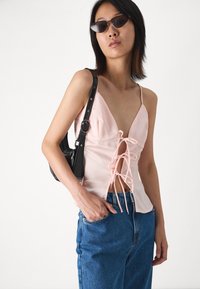 Rozā bez piedurknēm topiņš ar aukliņu priekšējā daļā, izgatavots no gludas auduma. Apvienots ar augsto jostasvietu ziliem džinsiem un melnu pleca somu.