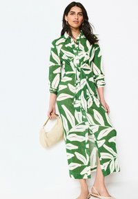 Femme portant une robe à motifs de feuilles verts et blancs, tenant un sac à main tissé beige et portant des chaussures à bride arrière beiges, sur fond blanc.