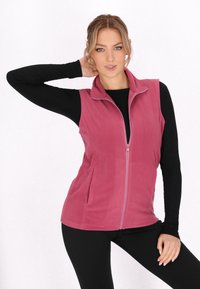 Gilet in pile rosa polveroso con colletto alto, chiusura con zip e due tasche laterali. Indossato sopra una maglietta nera a maniche lunghe e abbinato a leggings neri.