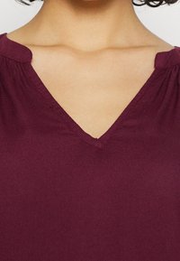 Top borgogna con scollo a V e maniche corte arricciate, realizzato in tessuto liscio. Presenta un delicato dettaglio di cucitura lungo il collo e le spalle.