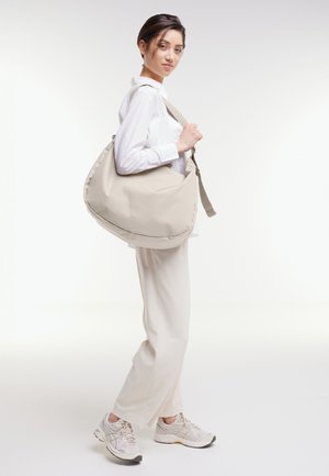 Femme en chemise blanche et pantalon portant un grand sac à bandoulière beige, chaussée de baskets assorties, se tenant sur un fond blanc.
