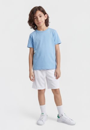 Ung dreng stående iført lyseblå T-shirt, hvide shorts, hvide strømper og hvide sneakers med grønne detaljer på ensfarvet baggrund.