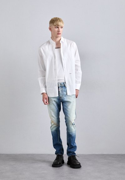 Chemise blanche à boutons, jeans bleu clair avec des détails usés, chaussures noires, se tenant contre un fond neutre.