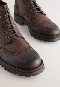 Braune Leder-Stiefeletten mit Flügelspitzenmuster, Perforationen an der Spitze und robuster schwarzer Gummisohle. Mit braunen Schnürsenkeln und Ziernähten.