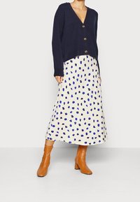 Navy knitted cardigan with a v-neck and wooden buttons, paired with a cream skirt featuring blue and black polka dots, complemented by brown heeled boots.