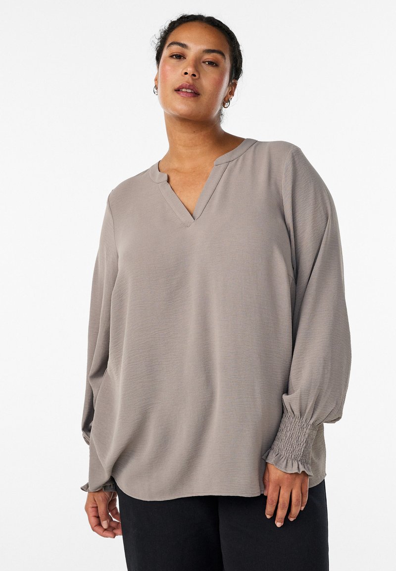 Žena oblečená v voľnej svetlosivej blúzke s dlhými rukávmi s nariasenými manžetami a výstrihom do V, v kombinácii s čiernymi nohavicami na bielom pozadí.