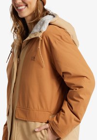 Brun och tan huvjacka med en fluffig innerfoder, med snördetaljer och framfickor. Slätt tyg med en avslappnad passform.