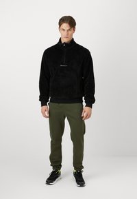 Sort fleece pullover med halv-zip krave, der har et broderet logo. Sammen med grønne joggers og sorte træningssko.
